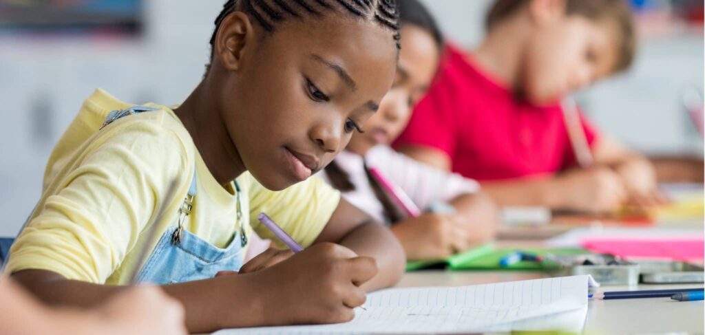 A little girl with braided hair wearing suspenders and a yellow t-shirt writes in a notebook at school. Two other children share the table with her.