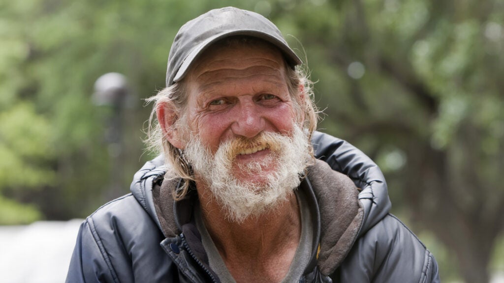 An older man wearing a cap and a hooded jacket smiles at the camera. He has longish gray hair and a gray beard.