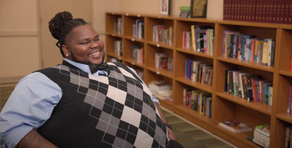 Mike, a graduate of Christopher Place Employment Academy, a program of Catholic Charities of Baltimore, is seated with a row of stocked bookshelves behind him and pictured with a big smile, wearing a blue oxford shirt, black bow tie and gray and white argyle sweater vest.