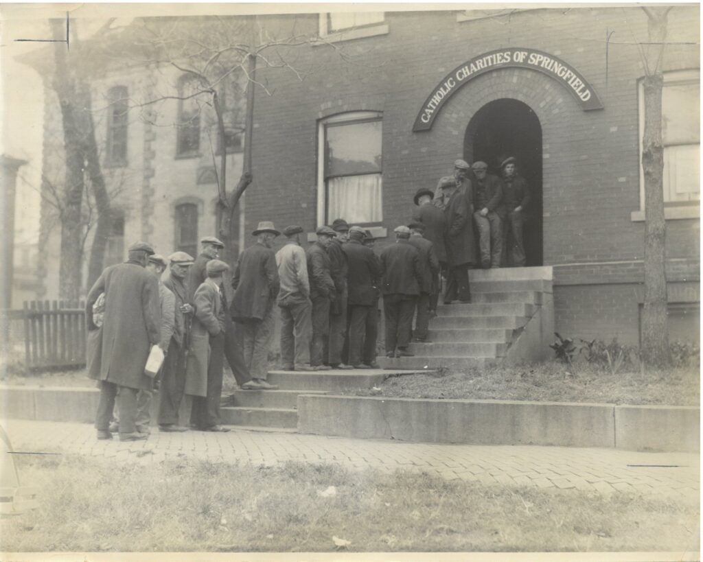 In a black and white photo, men line up outside a brick building with the words "Catholic Charities of Springfield" over the door, waiting for food.