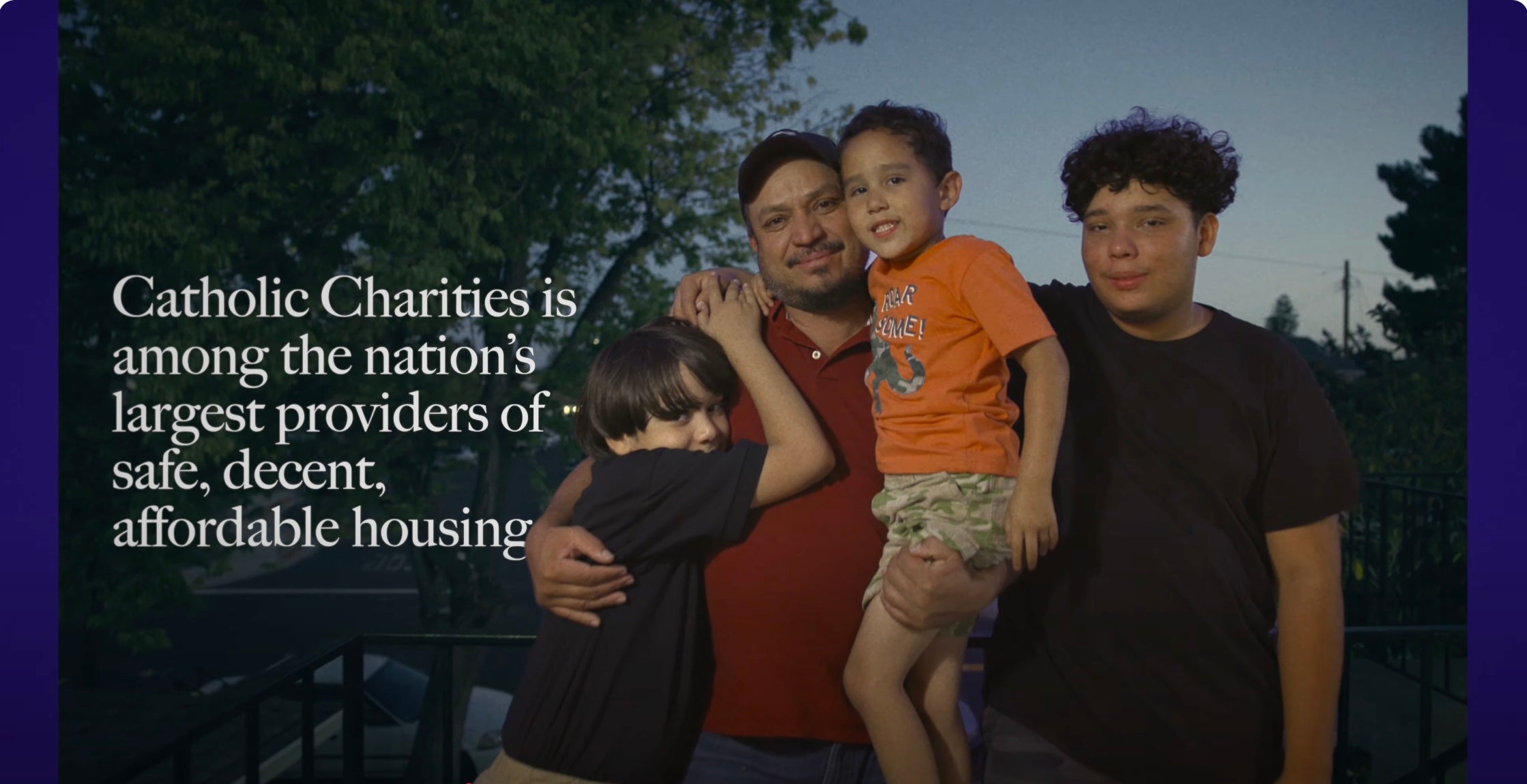 A father and his three children smile, arm in arm. The photo is a screenshot from a video and displays the text, "Catholic Charities is among the nation's largest providers of safe, decent, affordable housing"