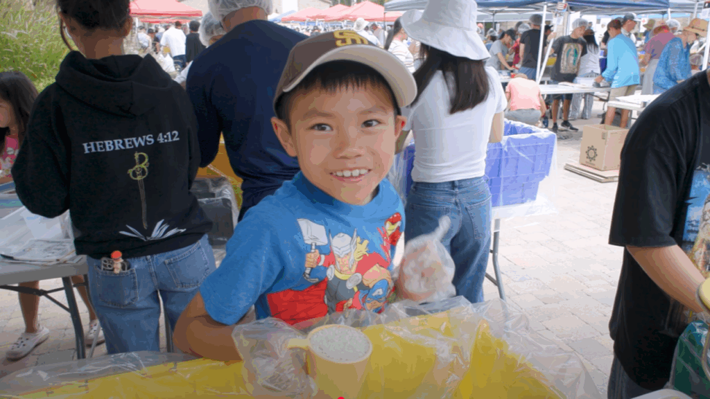 A little boy, smiling and holding a measuring cup full of rice, gives a thumbs up at the Million Meals Event, with other volunteers behind him packing food at long tables.