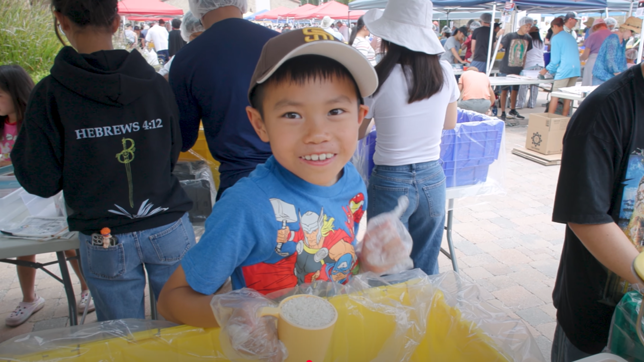 A little boy, smiling and holding a measuring cup full of rice, gives a thumbs up at the Million Meals Event, with other volunteers behind him packing food at long tables.